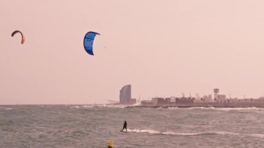 İki kişi okyanus sularının üzerinde paraşütle atlarken arka planda görünen bir şehir silueti var..