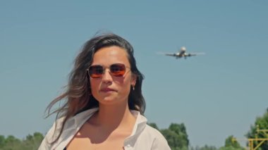 Güneş gözlüğü takan bir kadın güvenli bir şekilde park halindeki bir uçağın önünde duruyor..