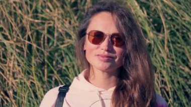 Güneş gözlüğü takan bir kadın uzun otların önünde duruyor..