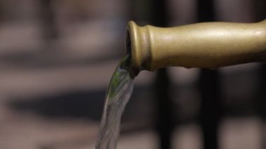Pirinç bir musluktan akan su, güneş ışığında parlıyor, yeşil ve yumuşak toprak tonlarıyla dolu huzurlu bir bahçe ortamı ile çevrili..