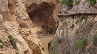 Turistler sarp kayalıklara oyulmuş dar bir geçitte dolaşırlar, dramatik bir kanyon manzarası ve aşağıdaki uzak tren yoluna bir göz atarlar..