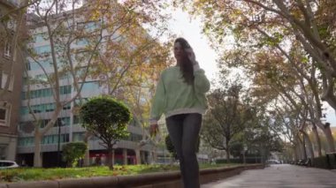 Bir kadın sonbahar renklerini gösteren ağaçlarla çevrili bir kaldırımda yürüyor, güneşli bir günde arka planda modern binalar var..