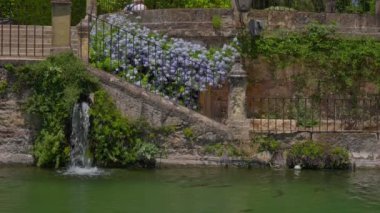 Serin bir şelale yemyeşil bir gölete akar yemyeşil bir bahçenin yanında çiçeklerle süslenir ve ziyaretçiler için huzurlu bir atmosfer yaratır..