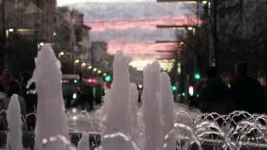 Gün batımının canlı renkleri gökyüzünü kalabalık bir caddenin üzerindeki yayalarla doldururken çeşmeden neşeyle su fışkırıyor. Akşam atmosferinin tadını çıkarıyorlar..