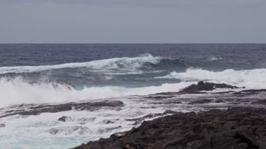 Güçlü okyanus dalgaları kıyı şeridi boyunca koyu kayalara çarpıp bulutlu bir gökyüzünün arka planına ve uzak bir ufka karşı beyaz köpük oluşturuyor..