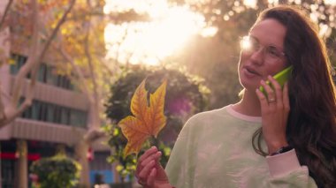Genç bir kadın telefonuyla konuşuyor, elinde canlı sarı bir sonbahar yaprağı var. Güneş arka planda batıyor, sahneyi aydınlatıyor..