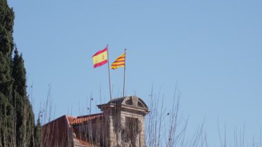İki bayrak gururla sallanıyor tarihi bir binanın tepesinde, İspanyol ve Katalan kimliklerini gösteren açık bir arkaplan ve çıplak dallara karşı.
