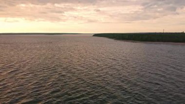 Yumuşak bir parıltı altında hafif dalgalar dalgalanır. Güneş sakin bir manzaranın arkasında batar. Gökyüzü ve su boyunca ılık renkler bırakır..