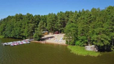 Ziyaretçiler sakin bir gölün yanındaki kumlu bir sahilde, parlak mavi gökyüzünün altında yükselen yeşil ağaçlarla dolu huzurlu bir günün tadını çıkarıyorlar..