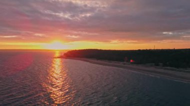 Golden hues blend with deep oranges as the sun sets over the calm waters, casting reflections on a serene beach and lush forest.