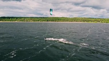 Yetenekli bir uçurtma ustası, rüzgarlı bir günde, verimli ormanlarla ve dramatik bulutlu gökyüzüyle çevrili dalgalarda sörf yapmanın heyecanını yaşar..