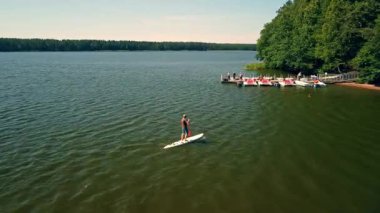 Bir insan durgun bir gölde süzülerek süzülür, yemyeşil ağaçlar ve uzaktaki canlı bir rıhtım tarafından çerçevelenir..
