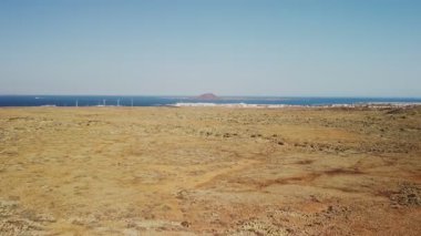 Uçsuz bucaksız karalar okyanusa doğru uzanıyor, açık mavi gökyüzünün altında küçük adalar yükselirken rüzgar türbinleri sergileniyor..