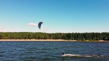 Yetenekli bir uçurtma sörfçüsü yumuşak dalgaların üzerinde zarif bir şekilde süzülür, rüzgarın gücünü yemyeşil ağaçların arka planına karşı kullanır..
