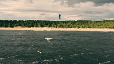 Yetenekli bir uçurtma sörfçüsü, rüzgarlı bir günde bereketli orman ve kumlu kıyılarla çevrili dramatik bulutların altındaki dalgalarda gezinir..