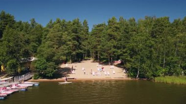 Canlı güneşlenenler sakin gölün kıyısındaki kumlu bir sahilde güneşlenirler. Etrafı gür ağaçlarla ve parlak mavi gökyüzünün altında canlı doğayla çevrilidir..