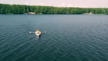 İki birey, berrak bir gökyüzünün altında canlı ağaçlar tarafından kucaklanan sakin bir gölde barışçıl bir kürek macerasının tadını çıkarıyor..