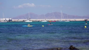 İki insan açık mavi sularda kürek çekmeyi seviyor, uzak dağlar ve parlak bir gökyüzünün altındaki canlı bir sahil cennetinde kayıklarla çevrili..