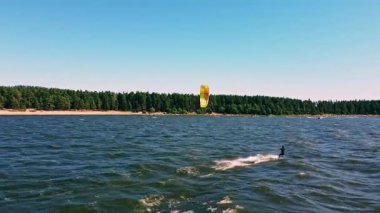 Yetenekli bir uçurtma sörfçüsü berrak mavi gölde zarif bir şekilde manevra yapar. Parlak, güneşli bir gökyüzünün altında yemyeşil ağaçlar tarafından çerçevelenir..