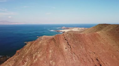 Canlı kızıl kayalıklar sakin mavi suların üzerinde dramatik bir şekilde yükselir, güneşli bir günde parlak mavi bir gökyüzü altında çarpıcı bir kıyı manzarası sergilerler..