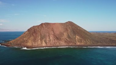 Engebeli bir volkanik ada berrak mavi gökyüzü ve parıldayan okyanus dalgalarına karşı dimdik ayakta duruyor..