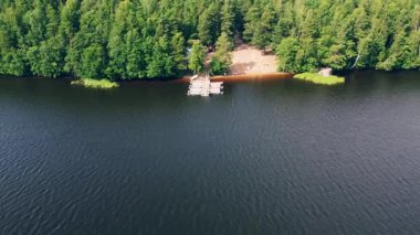 Huzurlu bir göl manzarası güneşli bir yaz gününde etrafı canlı yeşilliklerle çevrili, kumlu bir sahili olan ahşap bir rıhtımı sergiliyor..