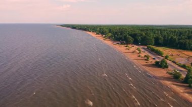 Güneşin altında sakin bir kıyı şeridi uzanır. Yumuşak dalgalar, canlı yeşillikler ve ağaçlarla bezenmiş kumlu kıyılara çarpar..