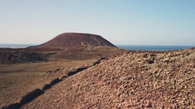 Engebeli bir arazinin üzerinde yükselen volkanik bir koninin nefes kesici görüntüsü, açık bir gökyüzünün altında okyanus görünüyor..