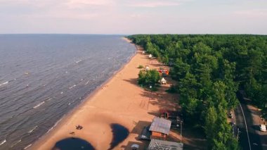Huzurlu bir göl kıyısı, yaz güneşinin tadını çıkaran ziyaretçilerle doludur. Arkaplanda yemyeşil ağaçlarla birlikte sular boyunca uzanır..
