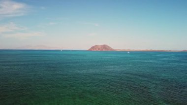 Kristal berrak sular, ışıl ışıl bir gökyüzünün altında, sakin bir adaya doğru uzanır, hafif dalgalar dalgalanır ve uzaktaki kayıklar huzur içinde süzülür..