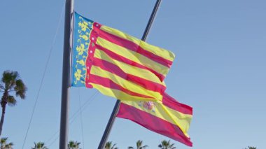 Rüzgarda dalgalanan parlak bayraklar açık mavi gökyüzüne karşı, palmiye ağaçları ile kıyı manzarası yakınındaki kültür ve mirası temsil ediyor..