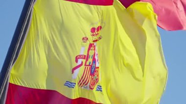 Canlı bir İspanyol bayrağı rüzgarda hafifçe dalgalanıyor, cesur renklerini ve karmaşık amblemini sergiliyor, ulusal gurur ve birliği simgeliyor..