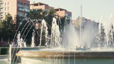 Su, güneşin altında zarif bir şekilde dans eder. Palmiye ağaçları ve kentsel binaların ortasındaki şelaleye hayran olan park ziyaretçilerini keyiflendirir..