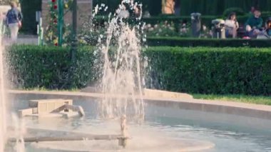 Bereketli yeşillik ve güneşli günün tadını çıkaran sakin park ziyaretçileriyle çevrili büyüleyici bir çeşmeden hoş bir su fışkırıyor..