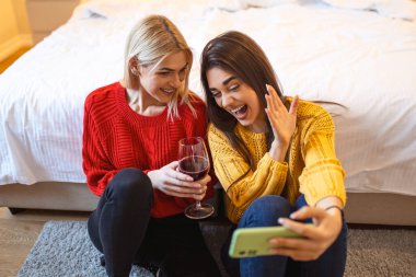 Evde yerde şarap içen arkadaşlar. Mutlu gülümseyen genç bayan arkadaşlar cep telefonundan fotoğraflara bakıyorlar. Kızlar eğleniyor. İki bayan arkadaş evde oturup bir bardak şarapla sohbet ediyorlar.