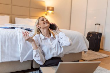 Otel odasından çalışan bir iş kadını. Güzel genç bir kadın cep telefonuyla konuşuyor. Yerde oturup iş seyahatinde eğlenmek.