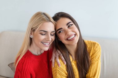 Sarışın ve esmer saçlı, kanepede örülü kazaklar giyen, sırtüstü oturmuş gülümseyen, sırtüstü kameraya bakıp koltuğa oturan iki güzel kadın.