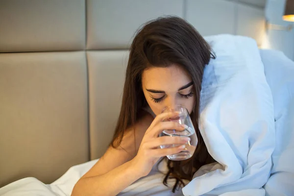 Young woman drinking glass of water in bed at night. Woman drinking a glass of water before going to sleep, she is lying in bed