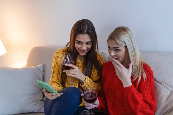 Friends with wine on the floor at home. Happy smiling young women friends looking at photos on mobile phone.