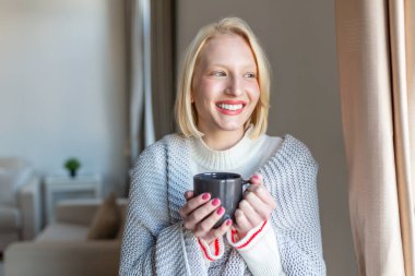 Sıcak bir battaniyeye sarılmış bir kadın elinde bir fincan kahve ya da çay içeceği ile pencerenin yanında duruyor.