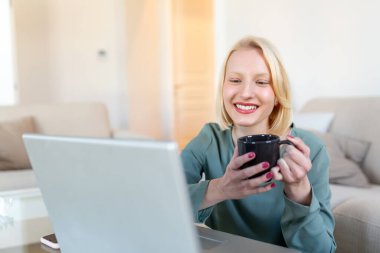 Positive young woman freelancer watching video in blog on netbook and having a coffee at comfortable home. Beautiful young business woman working on laptop.Freelancer connecting to internet