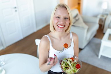 Portrait of a happy playful girl eating fresh salad from a bowl in her kitchen. Beautiful fit woman eating healthy salad after fitness workout