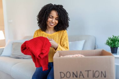 Happy African young woman sit on couch stuck clothes in donation box at home, caring biracial female volunteer put apparel in carton package, donate to needy people, reuse, recycle concept clipart