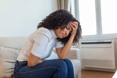 African American Woman in painful expression holding hands against belly suffering menstrual period pain, lying sad on home bed, having tummy cramp in female health concept