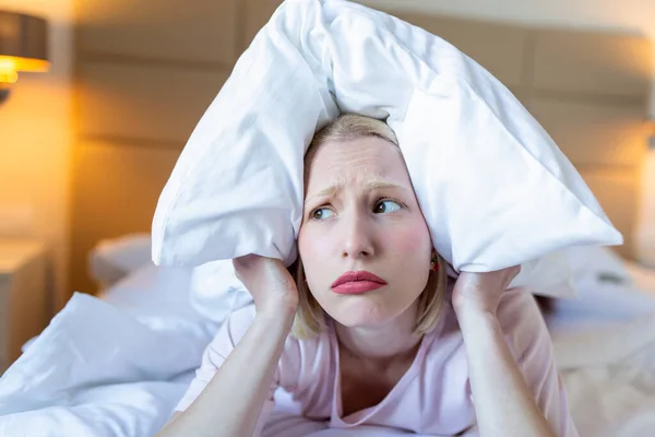 Angry woman suffering for neighbour noise lying on a bed in the night, covering ears with pillow because of noise.