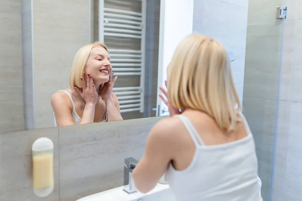 Cheerful young woman using cotton pad while looking in mirror. Happy smiling beautiful girl cleaning skin with cotton pad. Beauty natural woman looking in mirror while cleansing skin face