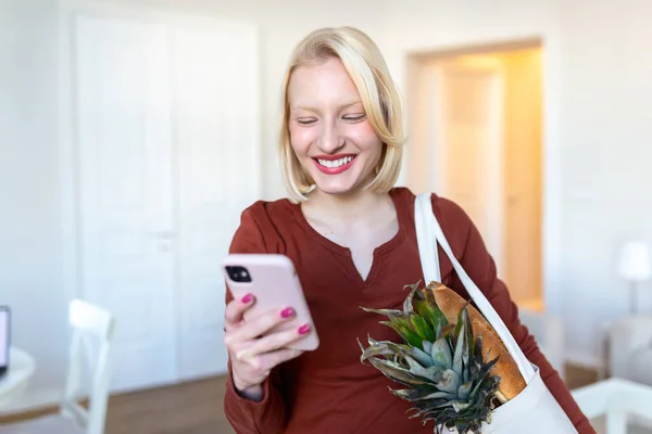 Pretty blond woman arriving home carrying groceries in a eco friendly tote bag using her smart mobile phone - Multi tasking