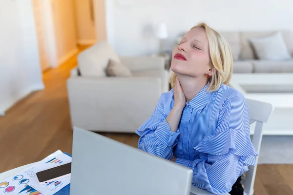 Portrait of young stressed woman sitting at home office desk in front of laptop, touching aching neck with pained expression, suffering from neck pain after working on laptop