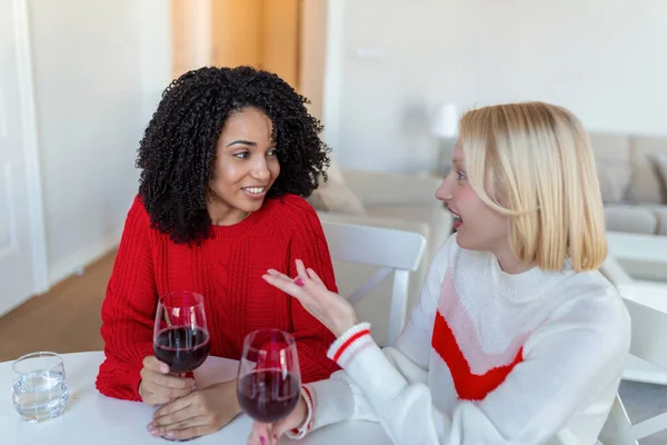 Women sitting on sofa laughing in a cozy loft apartment with wine. Two Female Friends Relaxing On Sofa At Home With Glass Of Wine Talking Together.