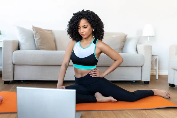 An adult African woman does yoga and strength training exercises on a mat in her living room. She follows an online exercise course video on her laptop.